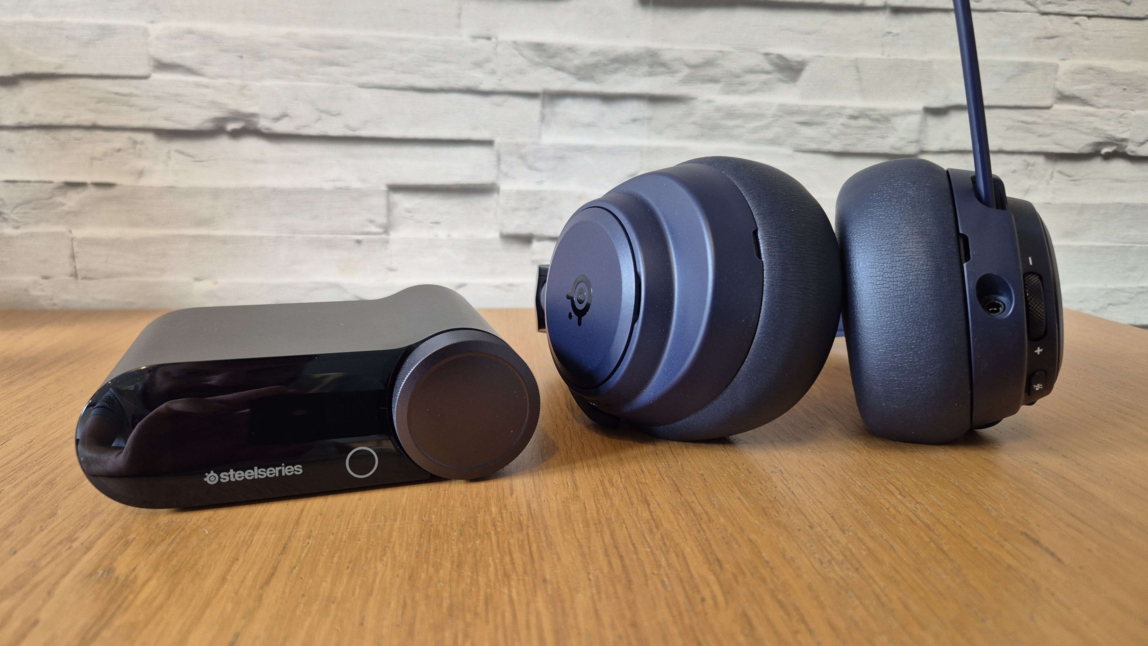 The SteelSeries Arctis Nova Pro Omni and it's GameHub in front on a white brick background and on a wooden surface.