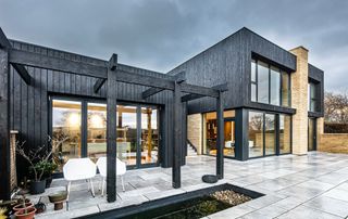 black clad modern home seen from rear with large black pergola structure and grey patio paving