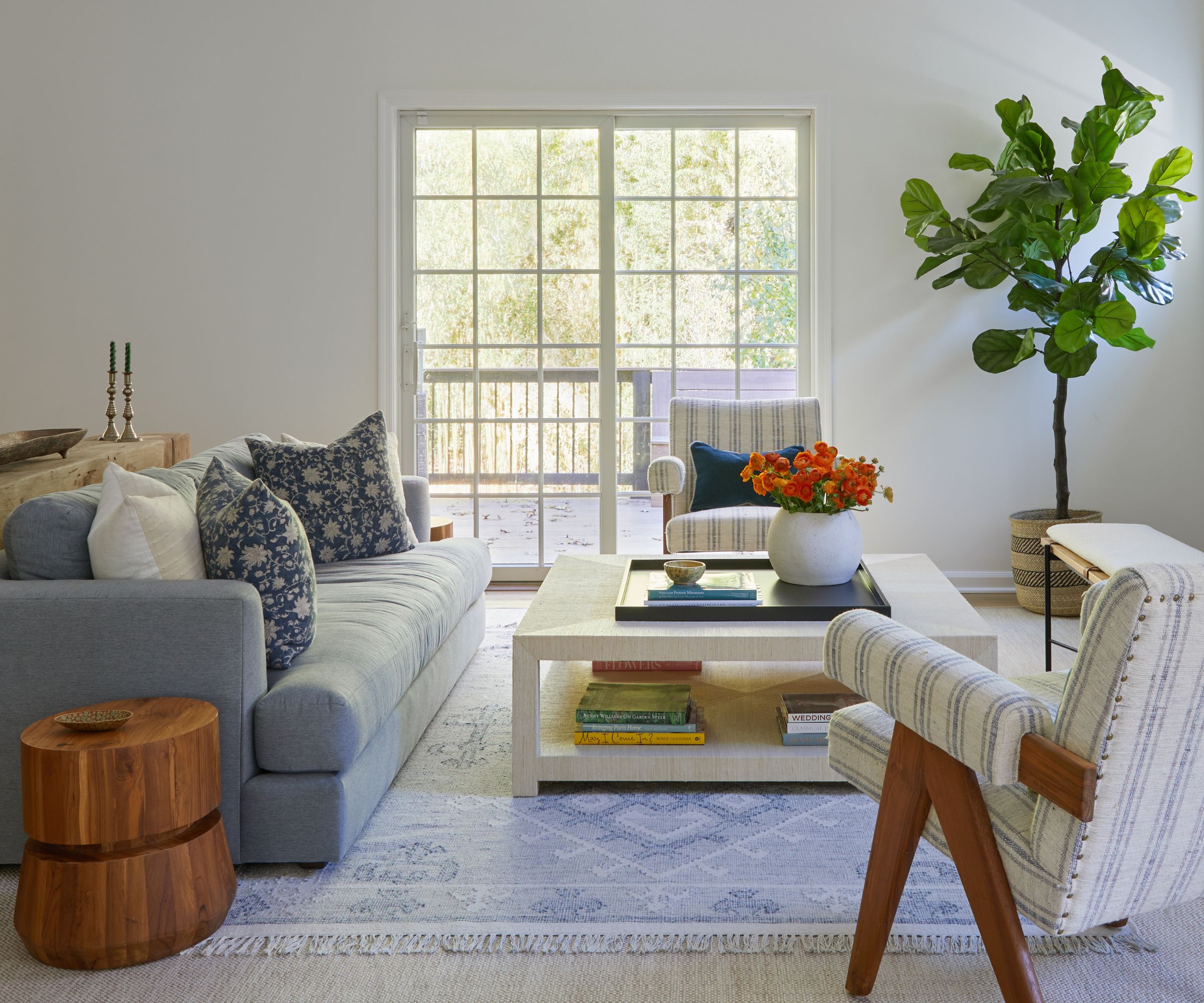 A coastal living room with white walls, glass doors leading to a balcony, and a pale blue sofa. A light blue patterned rug, a white-and-blue striped armchair, and a low white coffee table.