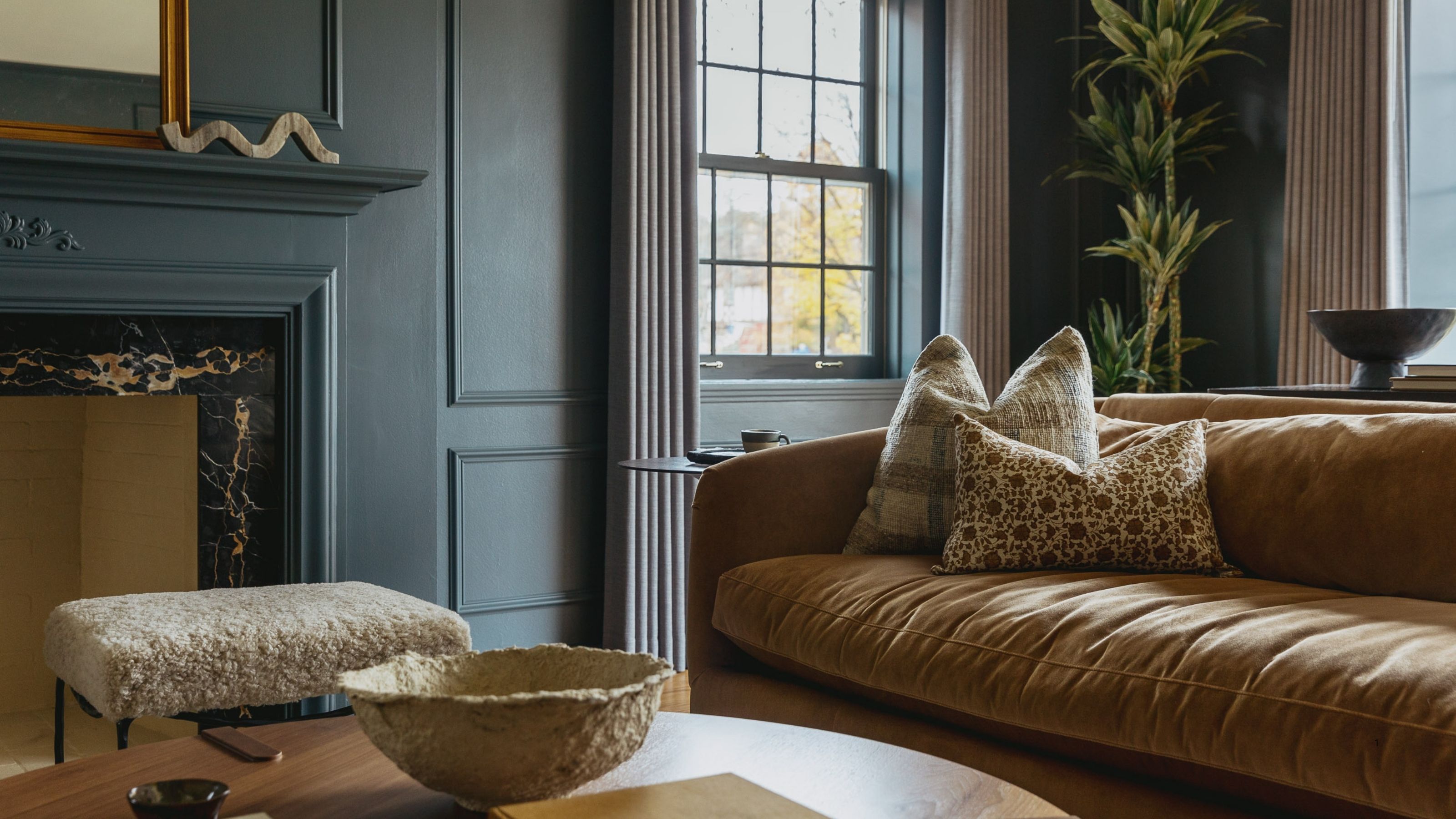 a dark blue living room with multiple windows, a fireplace, a wooden coffee table, and a tan, velvet sofa with throw pillows on it.