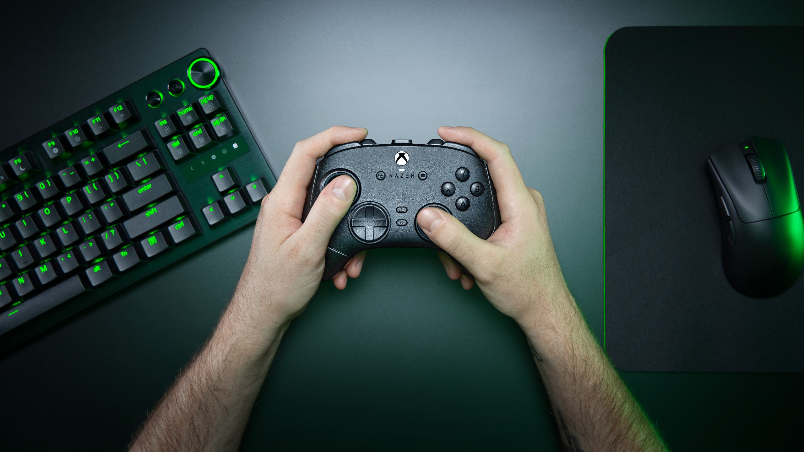 A lifestyle shot of the Razer Wolverine V4 Pro controller being held next to a Razer keyboard and mouse over a dark table.