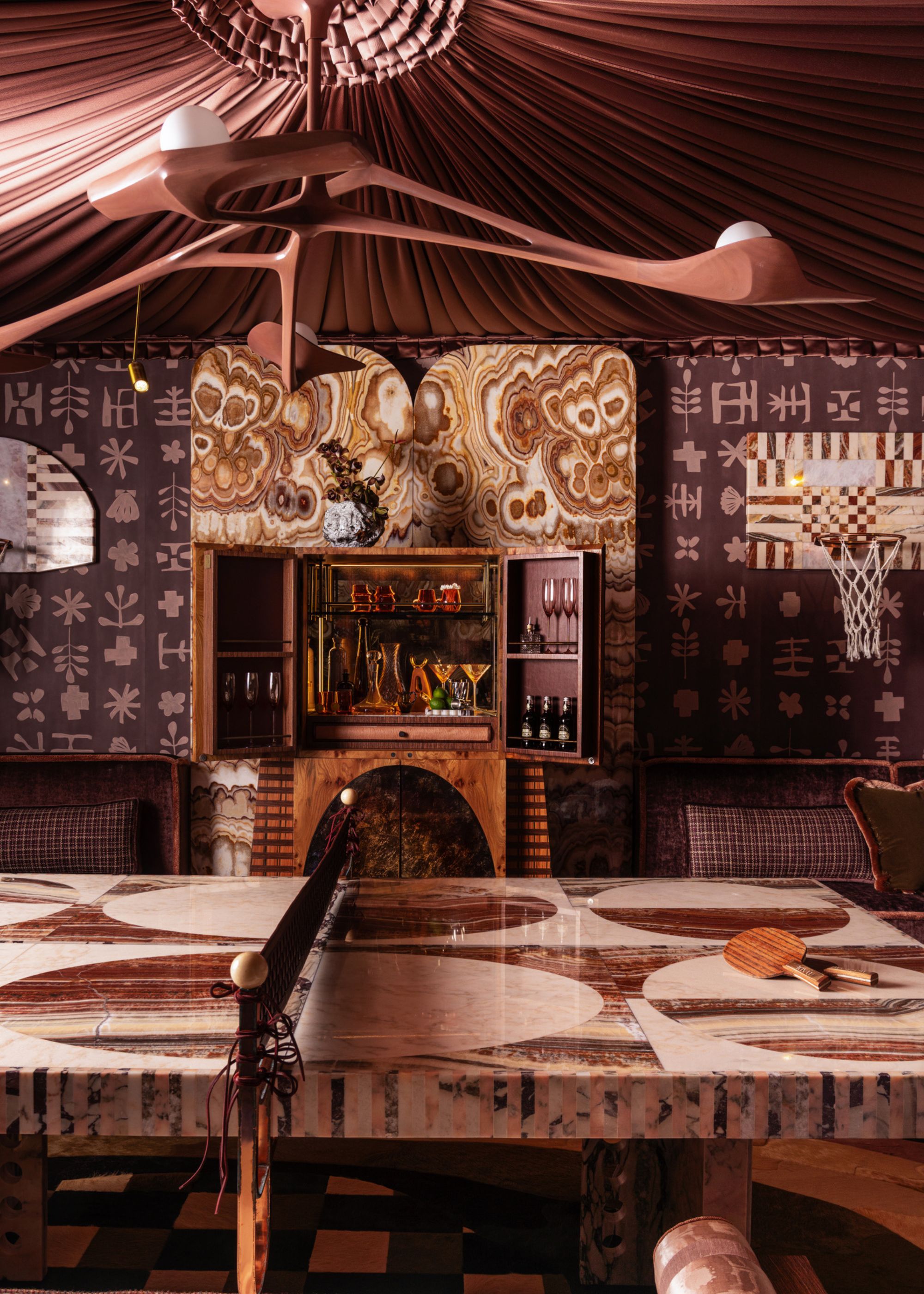 A moody and maximal rec room with a stone ping pong table with a geometric design. On the walls, there are purple-patterned wall coverings and a basketball hoop. In the centre of the wall, there is a bar area and a tented ceiling made from purple fabric.