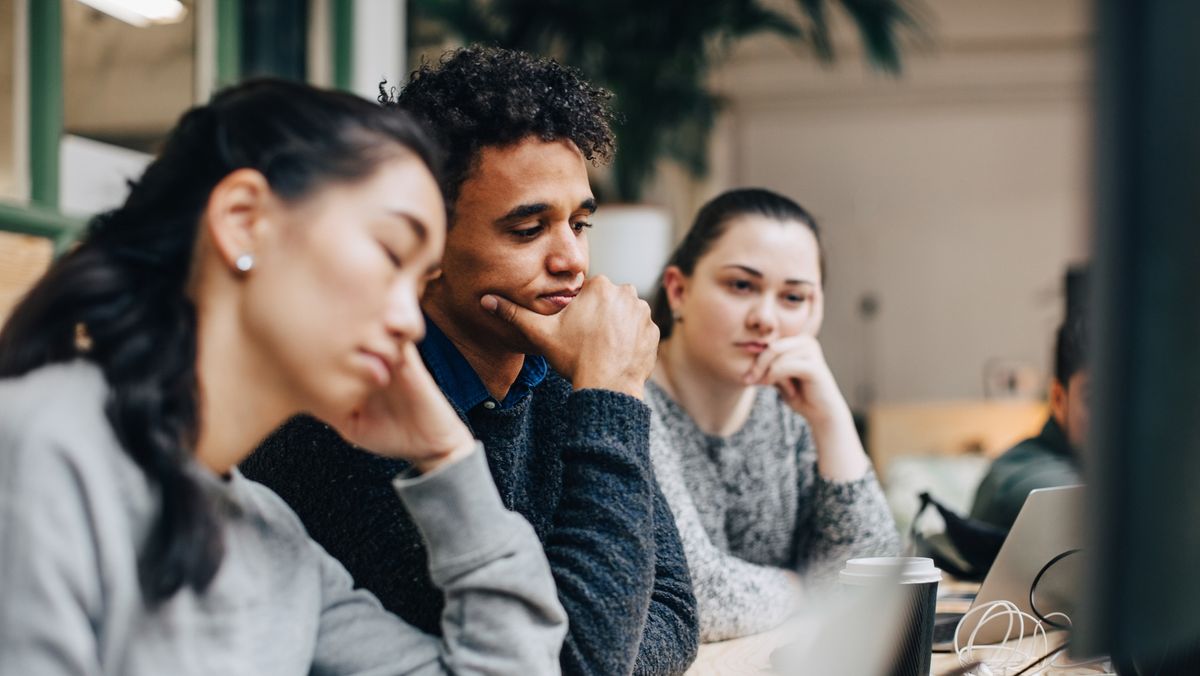 Three employees leaning on a large desk looking bored, to represent rust-out in the workplace. The three are shot from the chest-up.