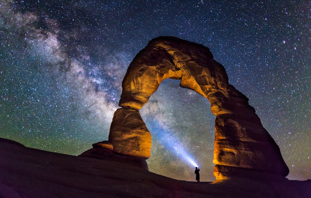 A person is standing in the middle of the Delicate Arch, Utah, USA, watching the milky way and lightning up the milky way with a flashlight beam.