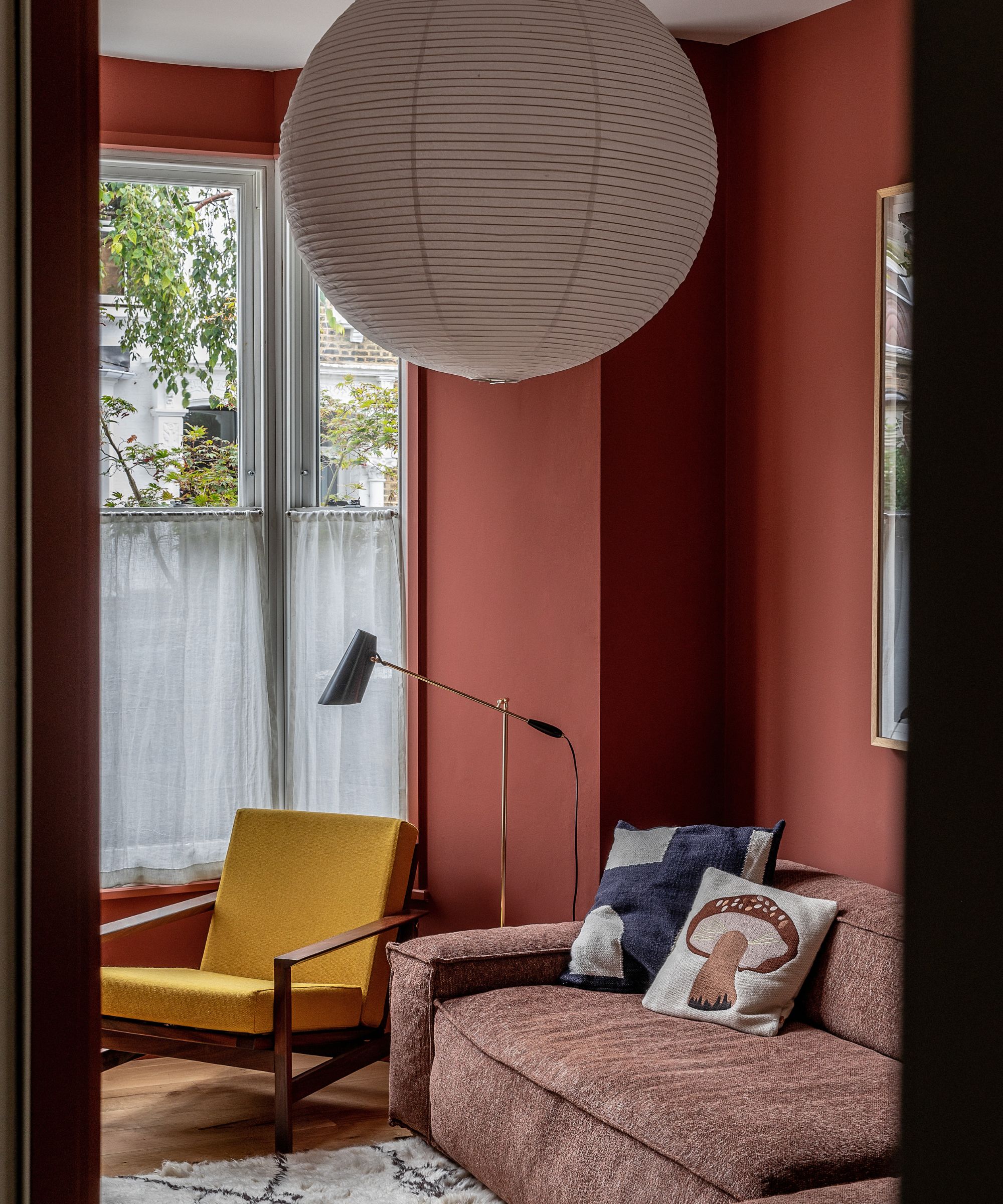 Minimal living room with earthy red painted walls, lantern pendant light, yellow midcentury chair, brown boxy sofa, Berber rug and gold floor lamp in the corner beside large bay window with cafe curtains