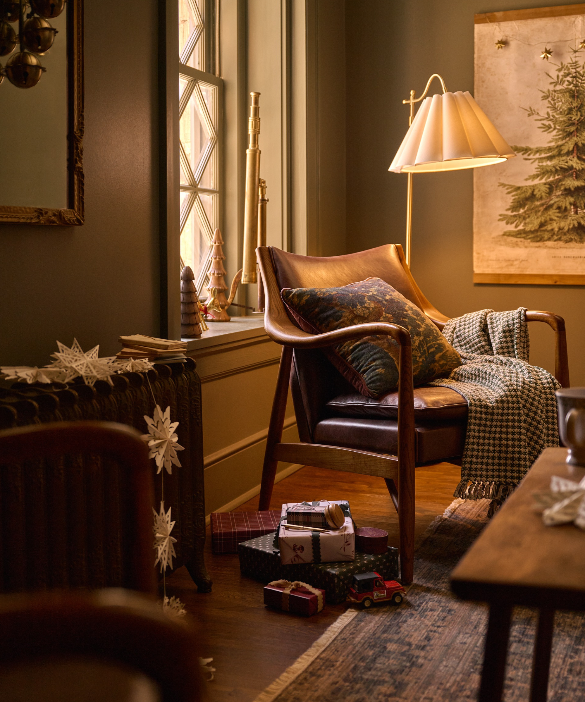 Cozy Christmas living room featuring paper cut-out snowflake garland, antique-style rug, wrapped presents, and a mid-century armchair with a tapestry-print throw pillow and plaid knit blanket.