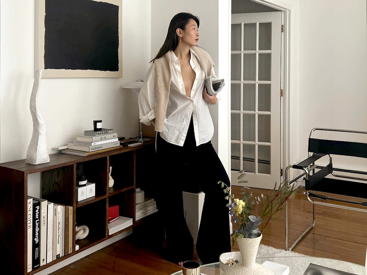 NYC fashion influencer Caroline Lin poses in her chic Manhattan living room wearing drop earrings, an open white button-down shirt, a neutral sweater over her shoulders, and black flared pants.