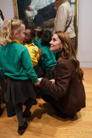 Kate Middleton wearing a brown blazer kneeling down talking to kids at an art museum