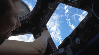 astronaut in front of a window facing earth. the astronaut is floating out the window at a barely visible piece of cylindrical equipment visible against the clouds