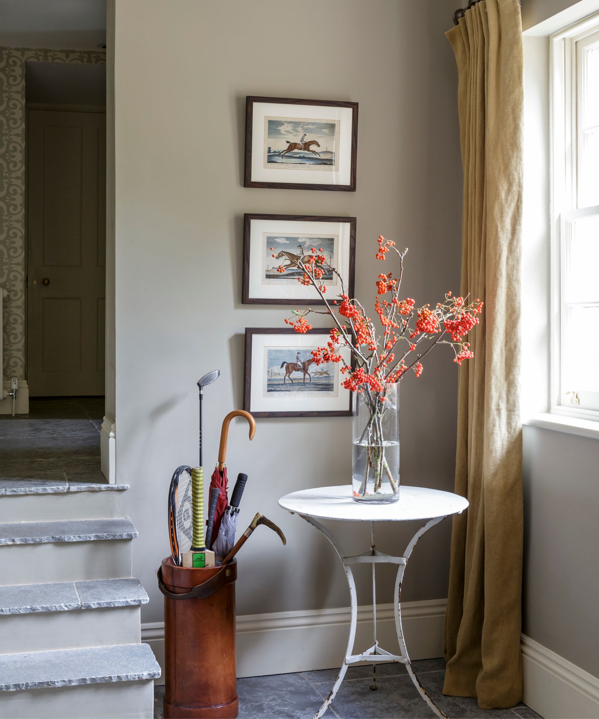 Entryway with slate floor and steps, a rustic white metal table holding a vase of autumnal branches, three equestrian prints hung on the wall, and an umbrella holder filled with umbrellas.