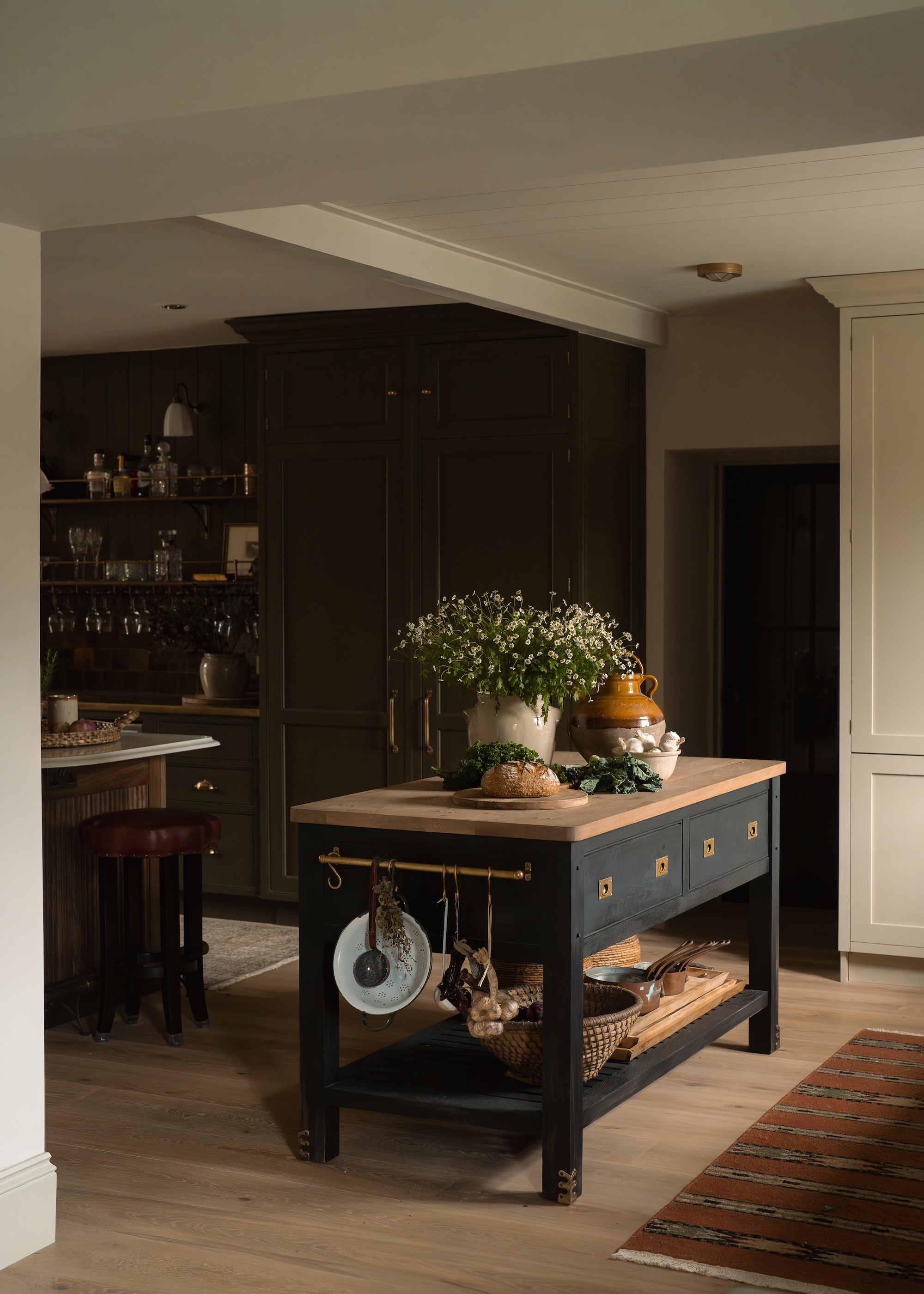 A dark black kitchen with a small rustic island in the middle. The island has drawers and a wooden countertop. On the counter is a vase and plants as well as other kitchen essentials.