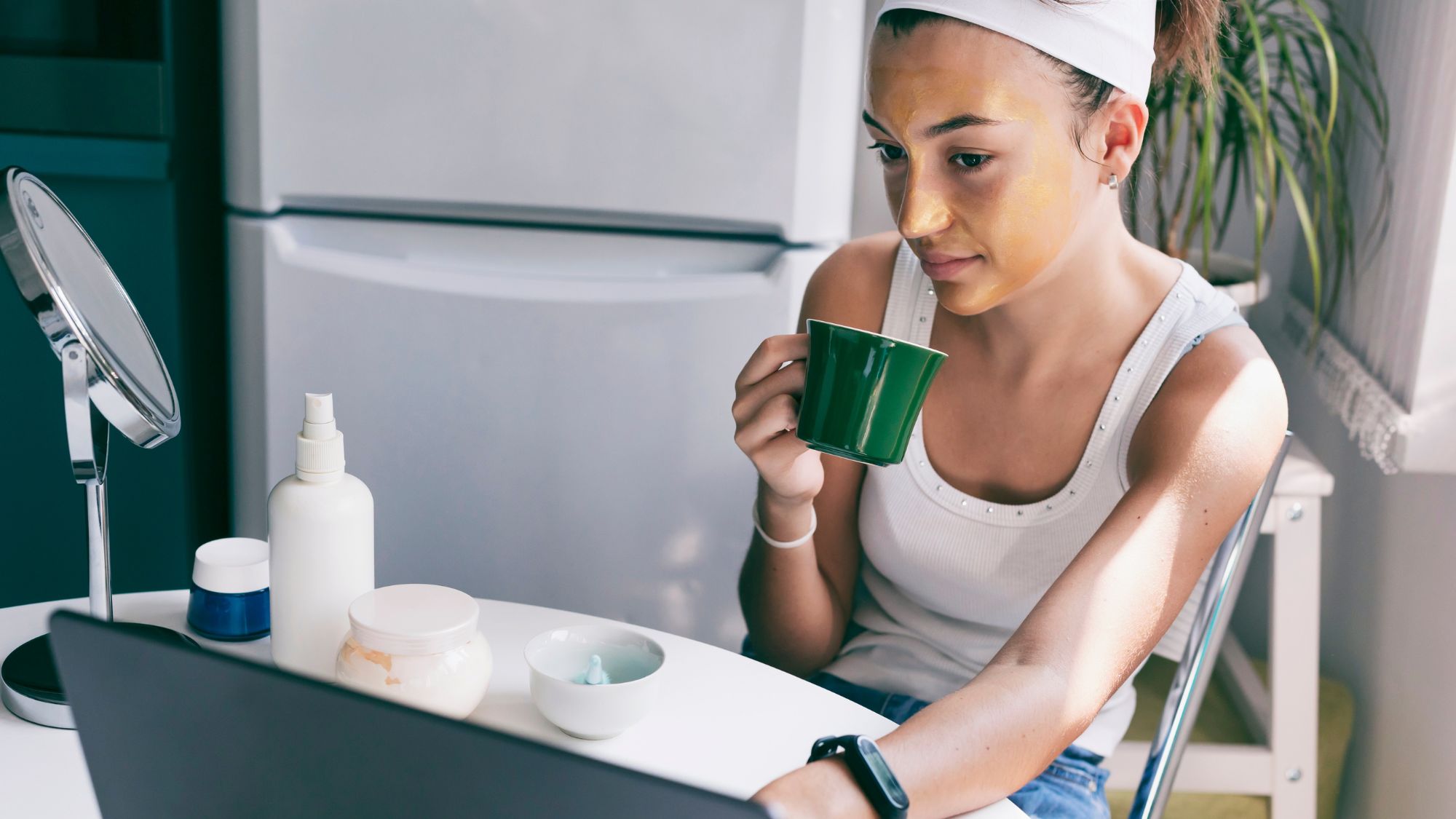 woman sat at her laptop with face mask on and beauty products around her
