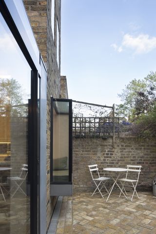 a glazed box window on the side of a brick terraced house, with a white dining set on the patio outside