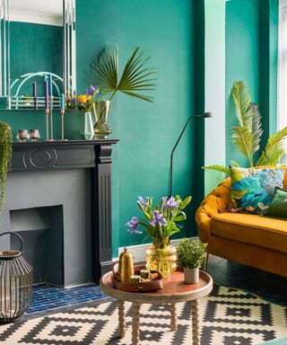 A living room with dark green walls, a black fireplace with decor on top, a yellow couch, a wooden coffee table with plants on top of it, and a black and white rug underneath