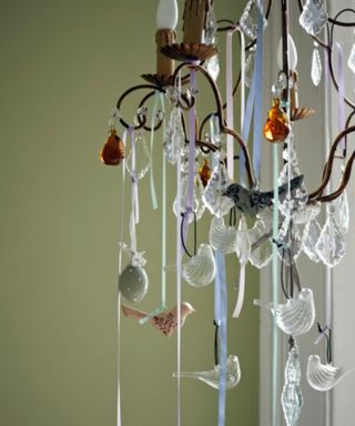 A collection of glass bird decorations hanging down with ribbons from a bronze chandelier, with a sage green wall in the background