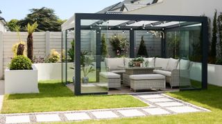 white painted horizontal timber fence behind low level white rendered wall with modern glass and black frame pergola over outdoor seating in garden area with grass and white paved path