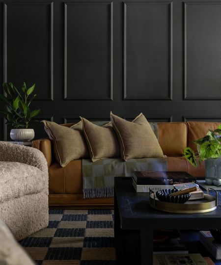 Living room with moody dark blue panelled walls, a rust linen sofa, linen cushions, a geometric throw, a checkered rug, two boucle armchairs, and a low coffee table with books, a plant, and a tray.
