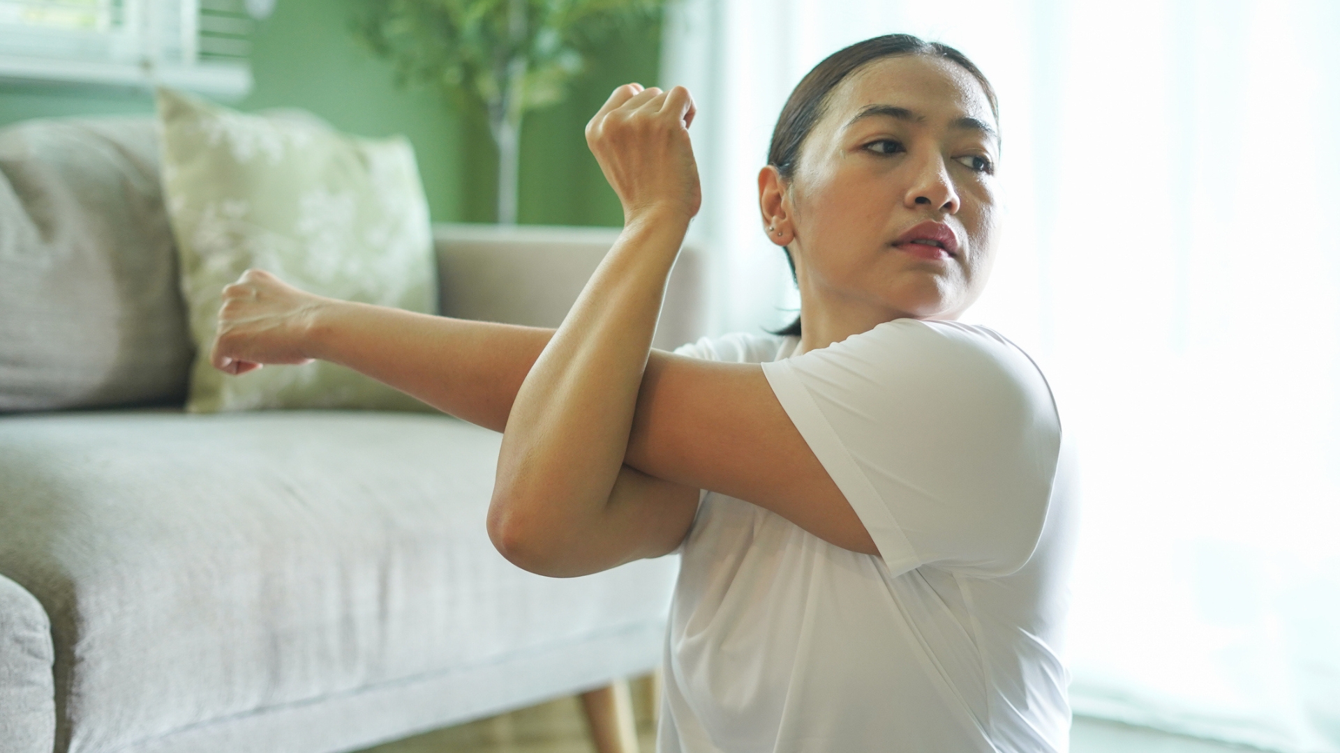 woman facing the camera pressing one arm across her body with the other arm. there's a sofa behind her. 