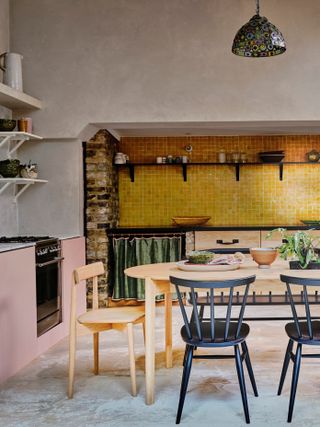 colorful kitchen dining space with shimmering gold backsplash, emerald green curtain, pink cabinets, black dining chairs, ercol table and chairs. open shelving, colorful pendant light