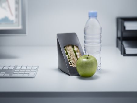 sandwich, bottled water, and green apple on an office desk with a monitor and paper tray in the background