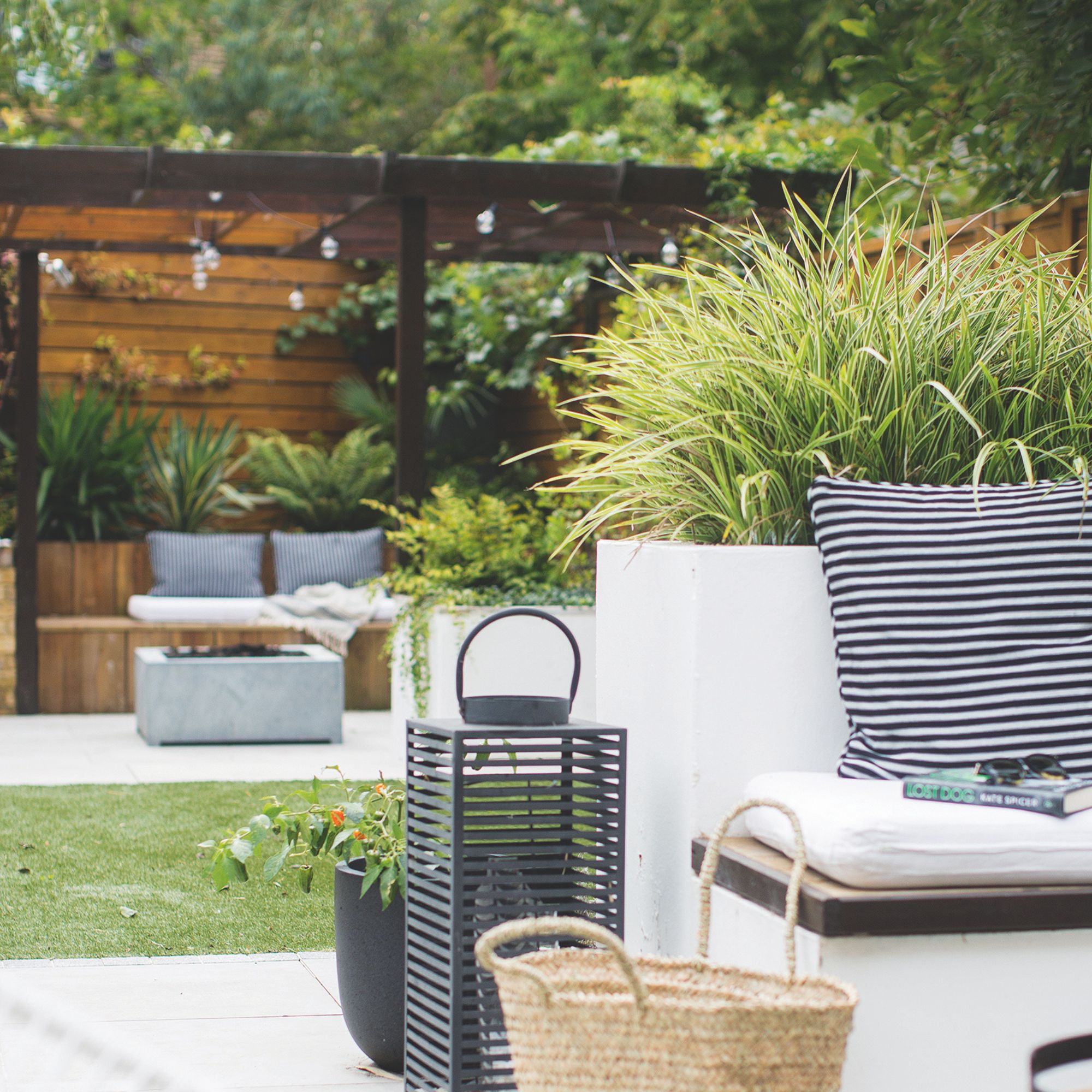 A garden seating area with a black lantern and striped cushion set against a backdrop of a dark brown pergola above wooden benches with cushions and string lights