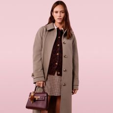 Model on a pink background holding a deep brown handbag wearing a grey collared trench coat, a brown plaid skirt and a brown cardigan sold at New Look