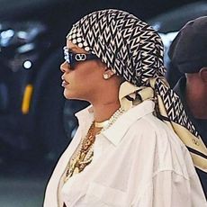 Rihanna wearing a silk scarf, black sunglasses, a white button down, blue jeans, and gold and pearl jewelry