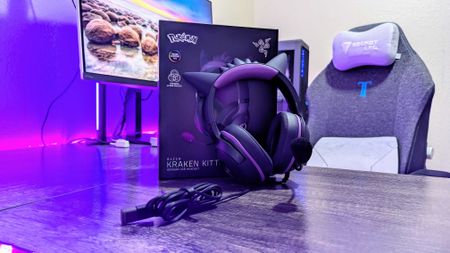 Purple-lit gaming setup featuring a boxed Razer Kraken Kitty headset with cat ears on a desk, alongside a gaming chair and monitor in the background.