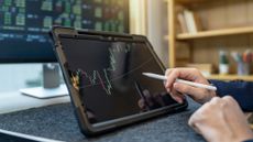 A man uses a digital pen to work on a tablet showing a trading graph, only his hands showing.