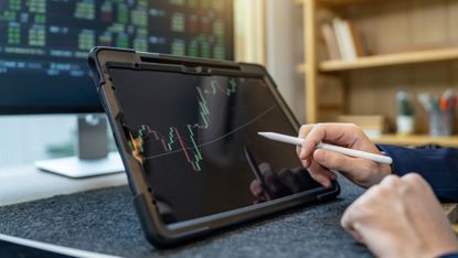 A man uses a digital pen to work on a tablet showing a trading graph, only his hands showing.