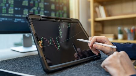 A man uses a digital pen to work on a tablet showing a trading graph, only his hands showing.