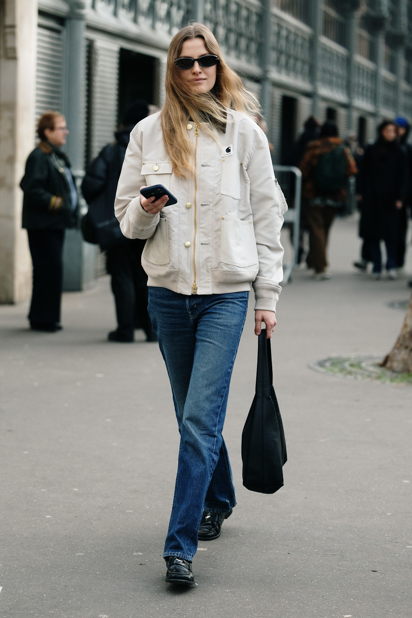 fashion week attendee wears chore jacket, sunglasses, jeans, and loafers