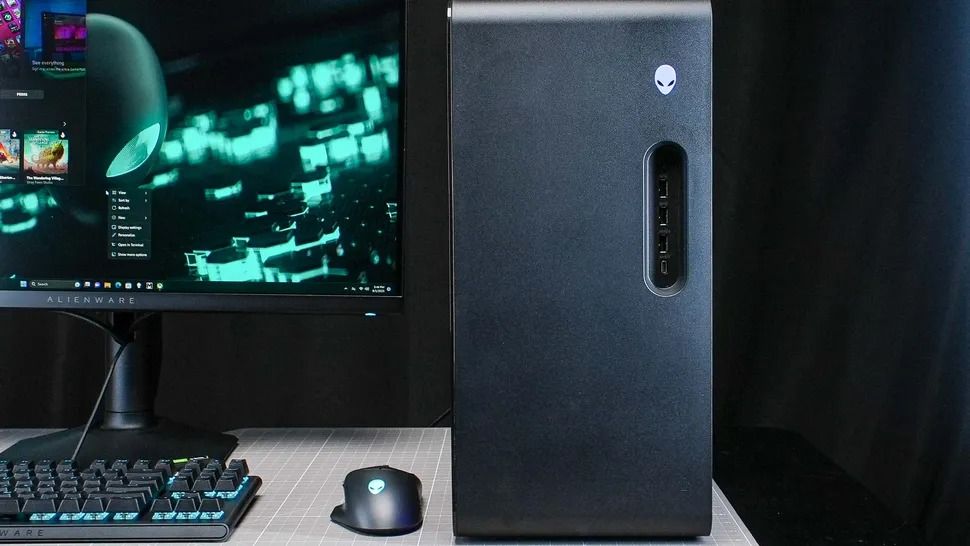 Alienware gaming desktop beside an ultrawide monitor showing glowing green code, with an RGB keyboard and mouse on a desk in a dark room.