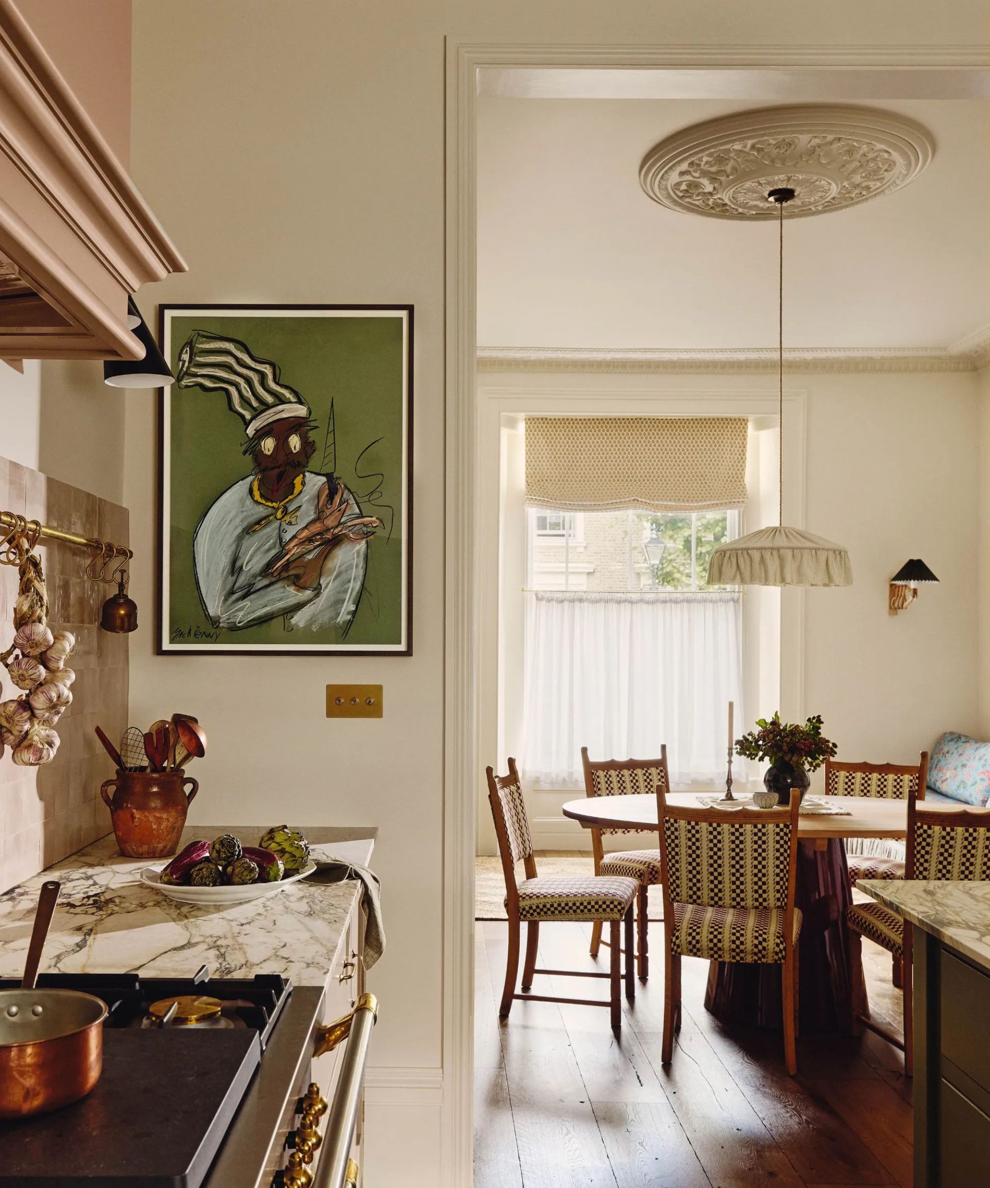 A corner of a traditional kitchen with marble countertops, abstract art on the wall, and a view into the adjoining dining space with a round wooden dining table and a large fabric pendant light above