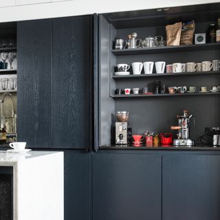 Dark grey kitchen cabinet with pocket doors open to show shelves full of mugs and a worktop coffee station set up