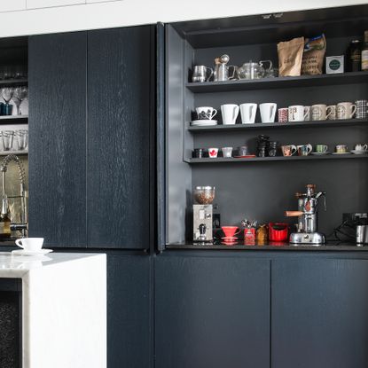 Dark grey kitchen cabinet with bi-fold doors open to show shelves full of mugs and a worktop coffee station set up