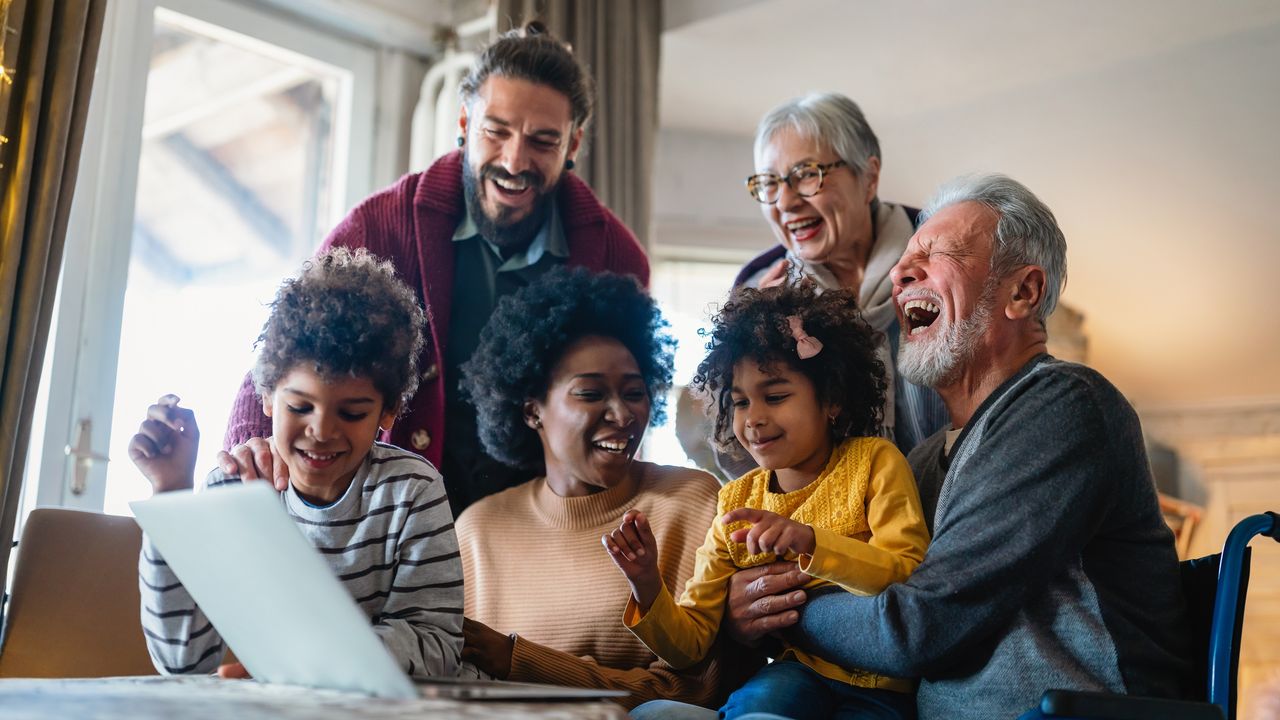 A multigenerational family laughs as they look at a laptop together on the dining room table.