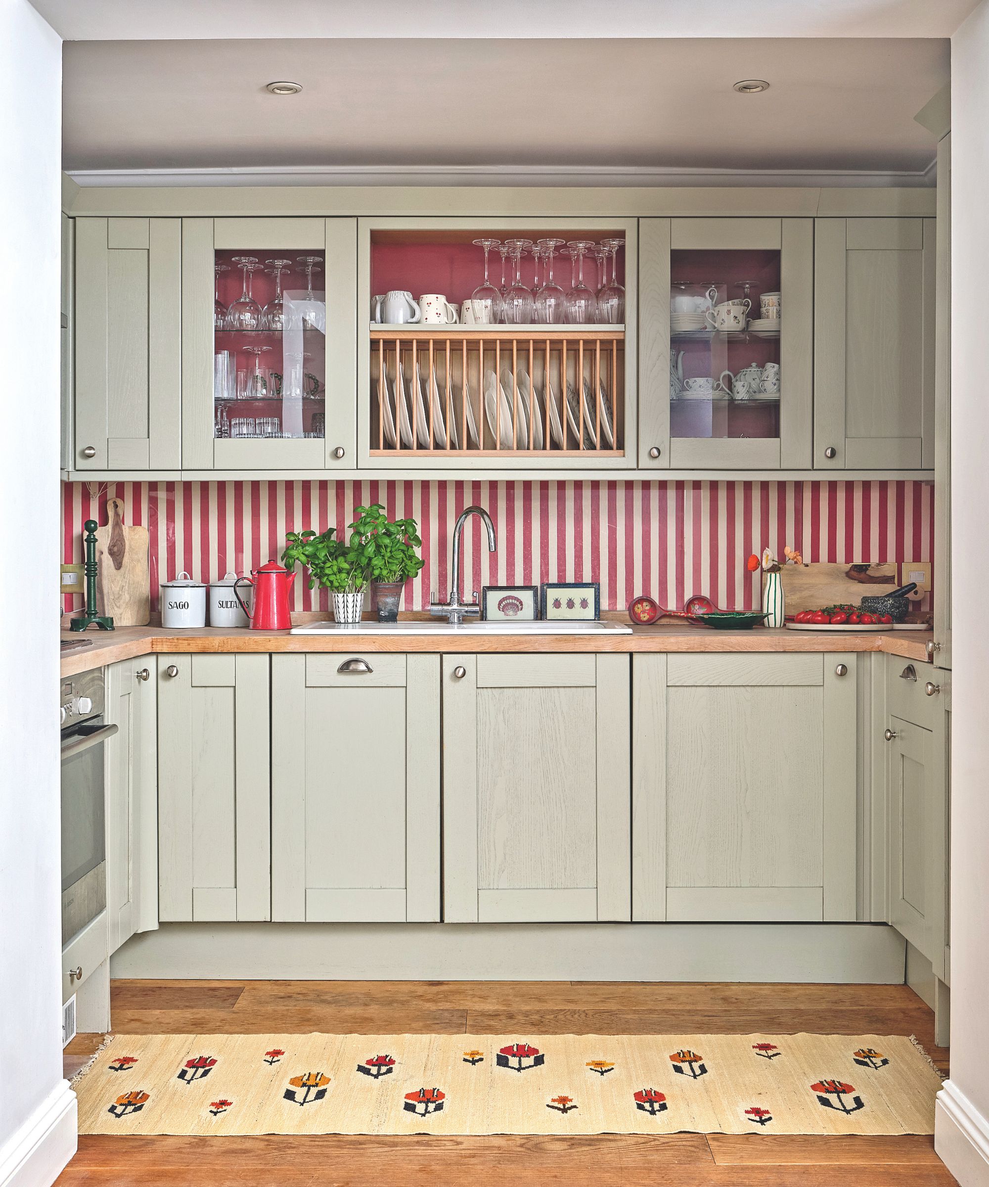 A pale sage green kitchen with a red and white vertical striped backsplash. The wooden countertops hold fresh herbs and a red teapot, with open shelving displaying glassware.