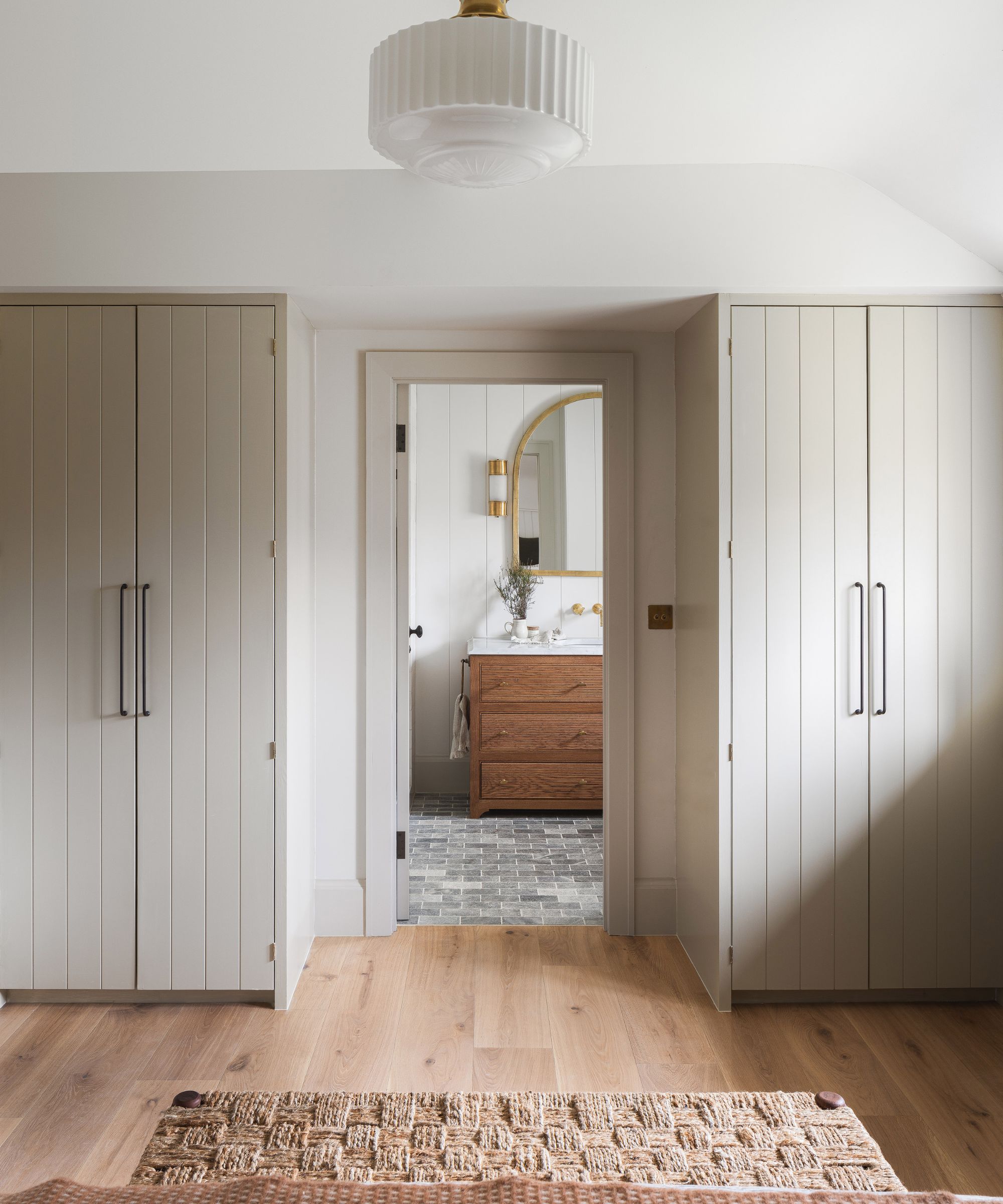 A symmetrical dressing area with two tall, cream built-in wardrobes flanking a central doorway. Pale wood flooring stretches across the room. Through the doorway, a wooden vanity with brass fixtures sits on grey brick-style tiles beneath an arched mirror.