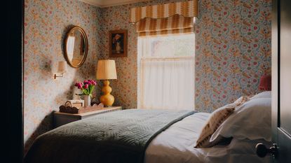 a floral wallpapered bedroom with yellow window blind and pelmet, and a small vignette with a dresser, lamp, scalloped tray, wall mirror, sconce and art