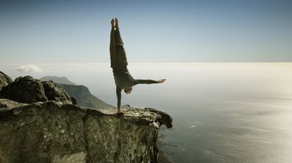 A man does a yoga pose, balancing on one hand on the edge of a cliff.