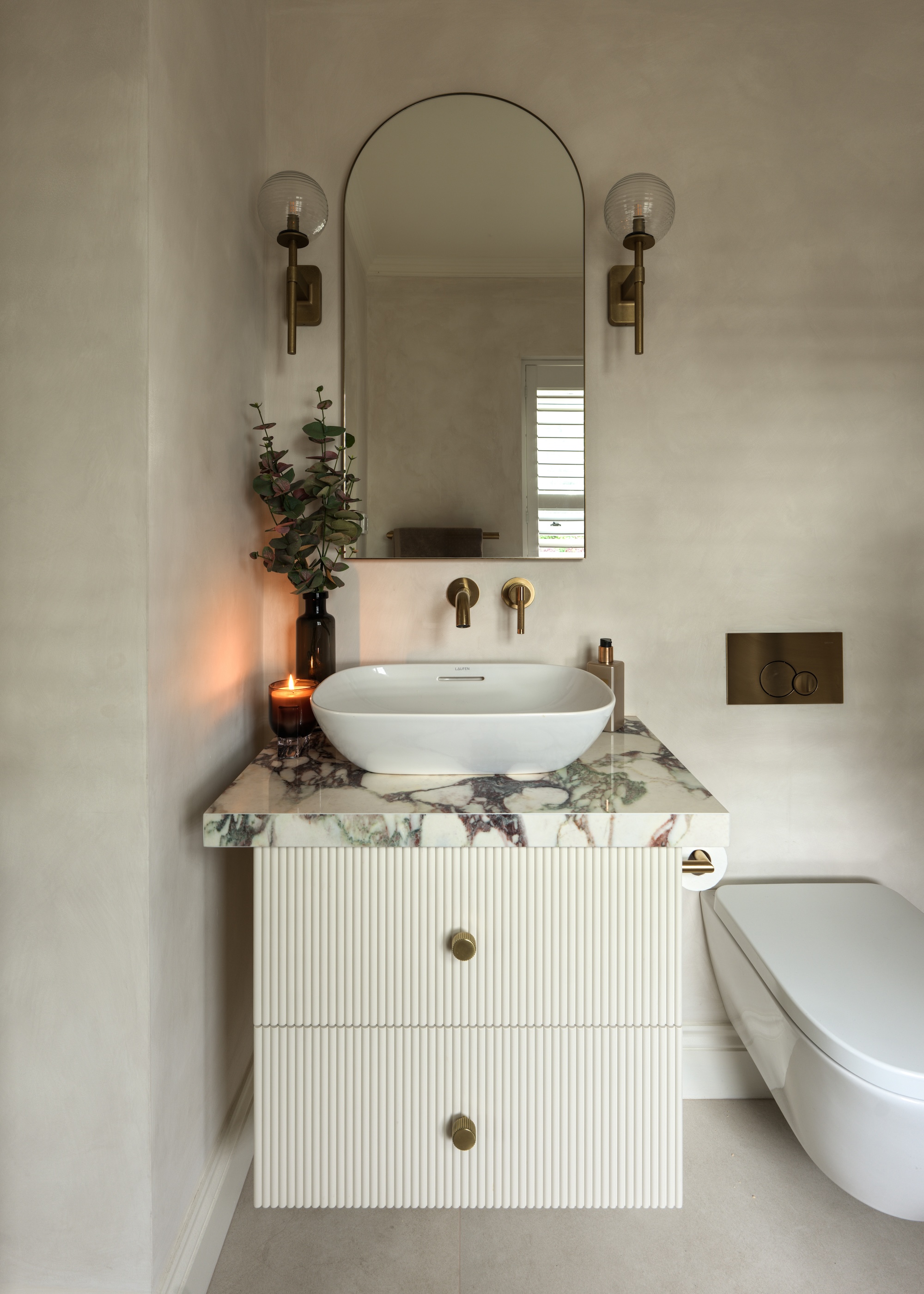 A small bathroom with a white vanity and marble countertop. Above the vanity is a mirror with wall lights on either side of it.