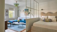 large bedroom with glass partition wall with murano glass pendant light, wavy striped headboard, white sheets, blue rug, with gray sofa with colorful cushion