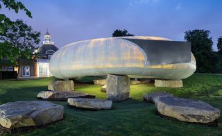 Alternative exterior view of the light grey, curved and illuminated 2014 Serpentine Pavilion by Smiljan Radic in the evening. The structure sits on large stones and is surrounded by greenery. There is also a building in the background