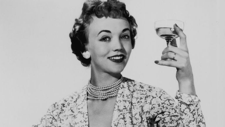 Lady with short curly hair wearing a floral dress and pearl necklace holding glass of alcohol