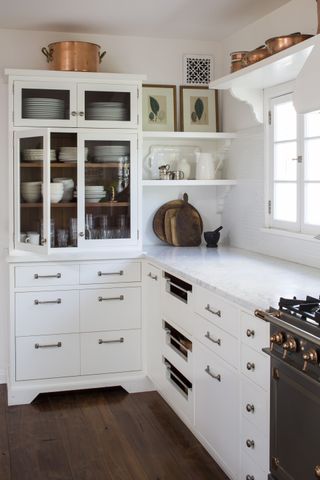 white kitchen with white cabinetry, marble countertops, glass fronted cabinet, open shelving, dark wood floor, copper pans, artwork