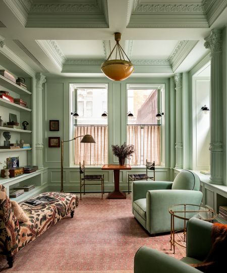 Living room with high ceilings and crown moulding painted celadon green, pink rug, floral daybed, green velvet armchairs and bookshelves with books and antiques