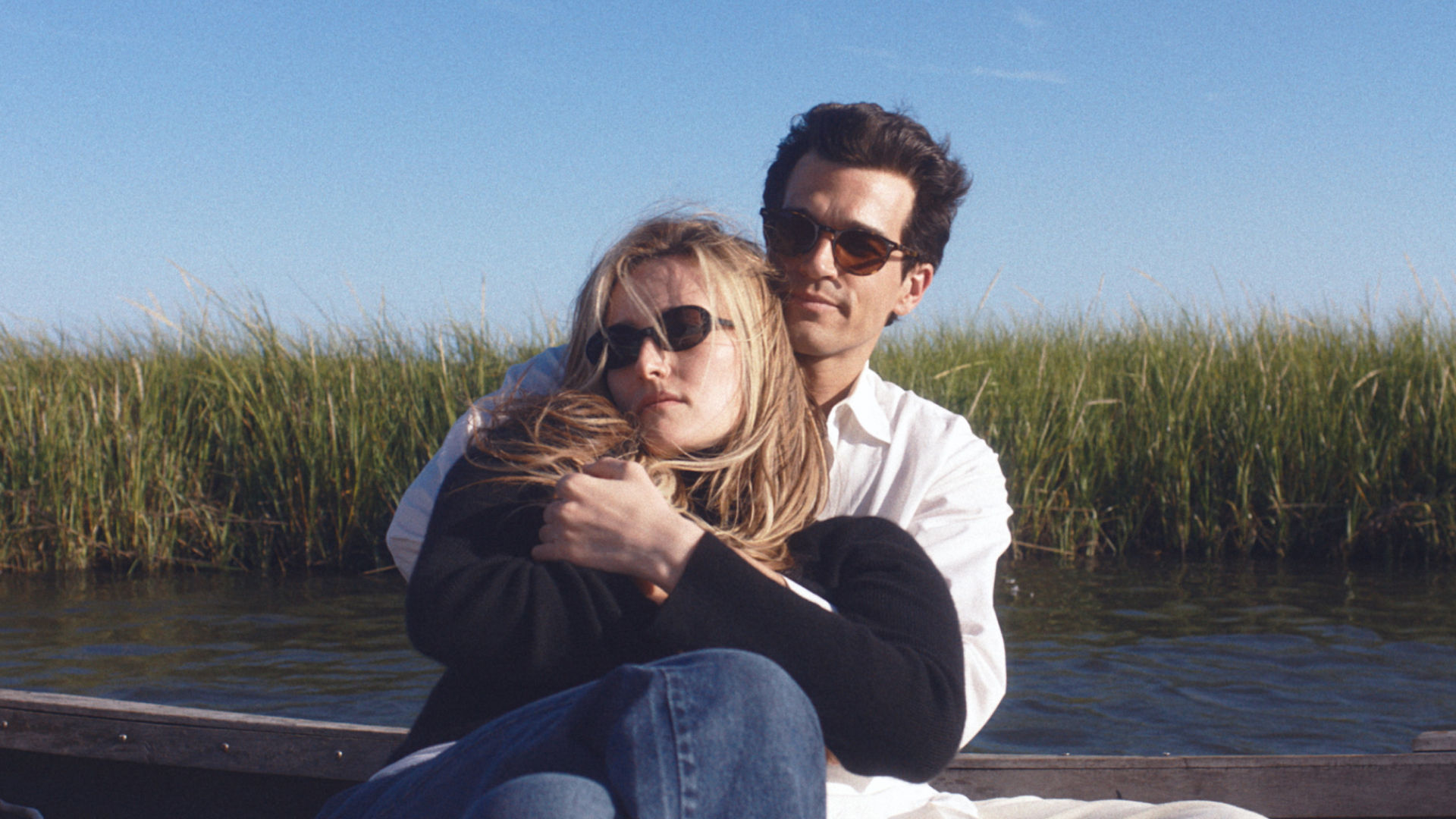 A blonde woman (Sarah Pidgeon as Carolyn Bassette Kennedy) and a brunette man (Paul Anthony Kelly as JFK Jr.), both wearing sunglasses, cuddle while sitting in a small boat on the water. A shoreline covered in reeds is seen in the background, in a still from FX's 'Love Story.'