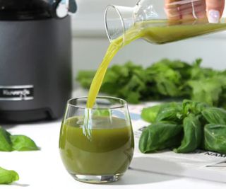 Green juice being poured into a glass with leaves and the Kuvings juicer in the background