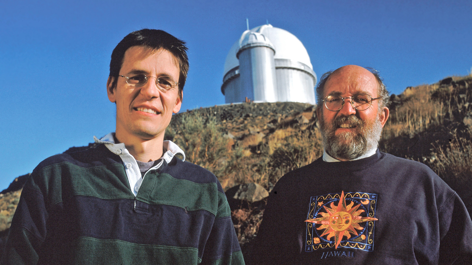 Two men stand next to each other with a large domed observatory in the background behind them